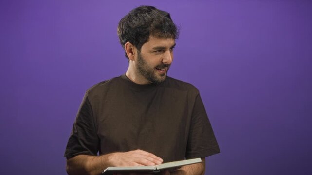 Man reads a book and flips pages with visible hand in purple studio setting while smiling slightly; quiet contemplation.