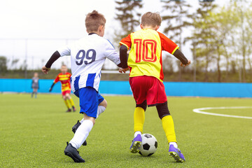 Fototapeta premium Junior Soccer Players in Blue and Yellow Kits Competing for Ball Possession During League Match - Youth Football Duel Between Number 99 and Number 10 on Turf