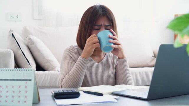 Sick Asian woman drinking hot water to soothe sore throat while working on laptop at home office. Concept of self-care during illness, flu symptoms, and remote work health management