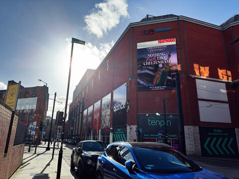 Sunlit urban street outside Printworks entertainment complex with Vue IMAX and Tenpin in Manchester, UK, showing cars, signage and red brick building facades