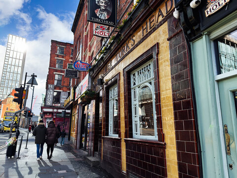 Pedestrians outside the Lower Turks Head pub on a busy Manchester, UK street with shops, buses, red brick buildings and classic pub facade