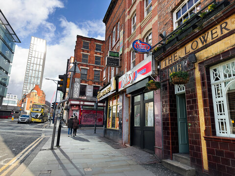 Street scene in Manchester, UK with traditional pub frontage, takeaway shops and a man standing outside Dixie Chicken on a busy urban sidewalk near double decker buses
