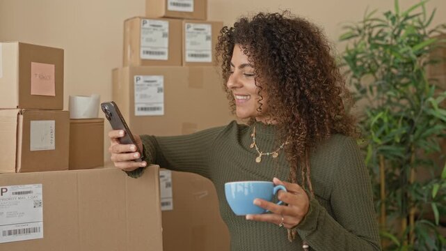 Woman holding smartphone with one hand and coffee mug in other by stacked parcels and shipping boxes inside a building; entrepreneurship optimism.