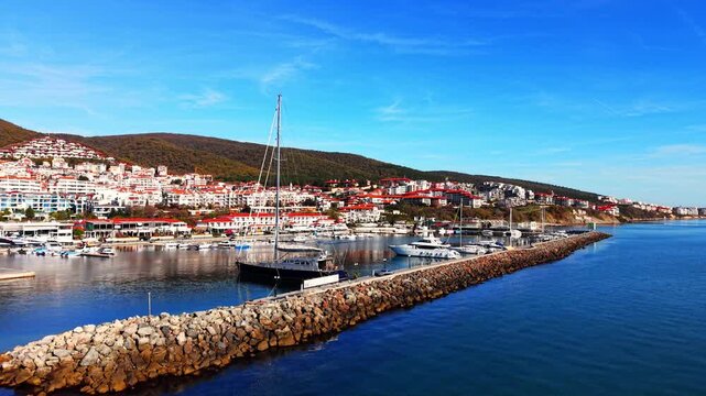 Aerial perspective of a yacht marina protected by a stone breakwater with a hillside resort town and clear blue sky. Yacht harbor and coastal view.