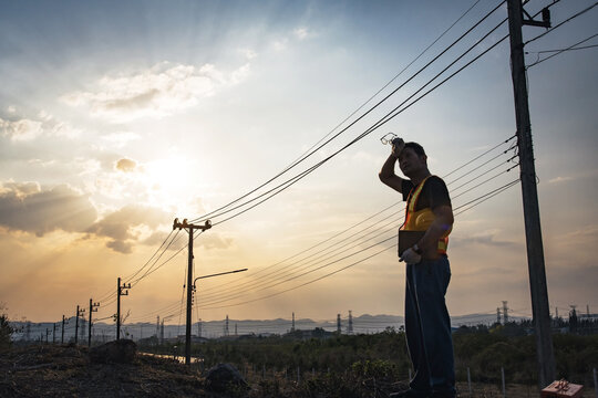 Male electrical engineer wipe sweat on his forehead, outdoor utility pole inspection during hot temperature in summer, silhouette sunset with sun flares, empower International Labour Day (May Day)