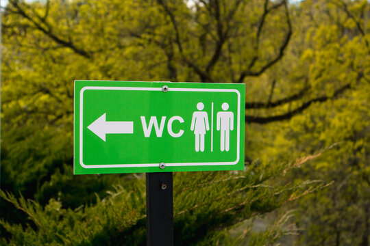 Bright green restroom sign with male and female icons pointing left in a park or natural outdoor setting.