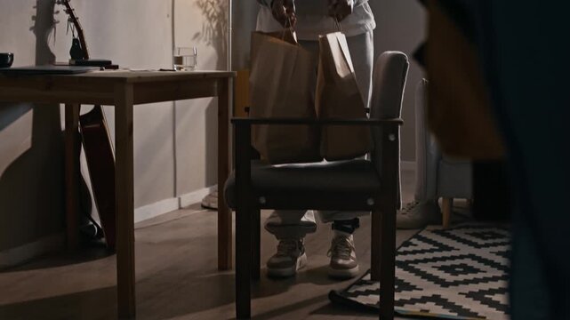 Tracking tilt up shot of Biracial woman under house arrest placing groceries from paper bags onto table while wearing ankle monitoring device at home