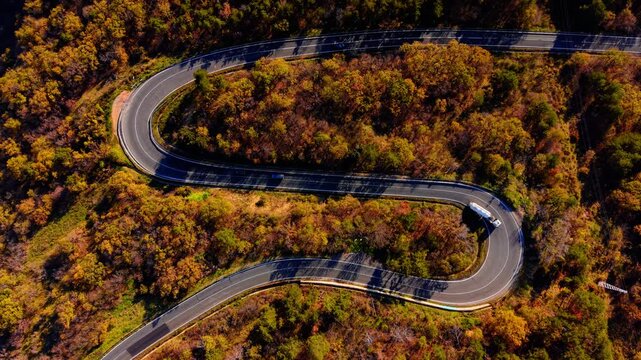 Aerial perspective of a winding asphalt road with cars driving through dense autumn forest. Top down view of serpentine road in autumn woods.