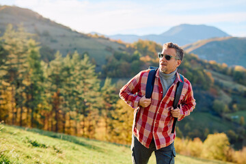 Fototapeta premium Middle-aged man with backpack and sunglasses hiking autumn mountains, enjoying sunlit forest vistas and freedom on a scenic getaway
