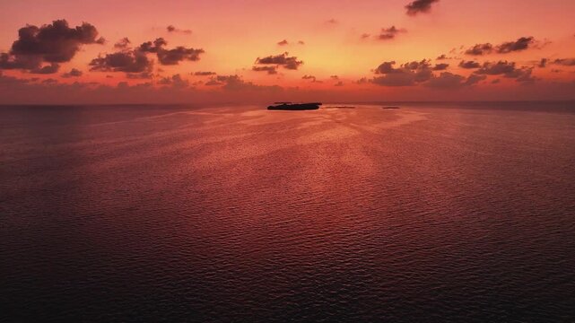 Aerial view of a vibrant sunset over the calm Indian Ocean with a distant island on the horizon in Goidhoo, Baa Atoll, Maldives.