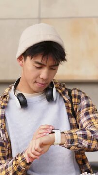 Vertical video: Raising left wrist, beanie-clad student checking white watch at plaza wall, tapping