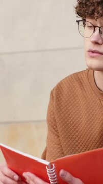Vertical video: Looking down at red notebook man with glasses in brown top reading notes on bench