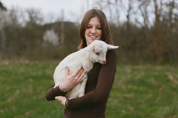 Naklejka premium A smiling woman outdoors holds a young goat kid in a grassy field, presenting a warm, gentle lifestyle moment. The scene emphasizes bonding, care, and natural companionship between human and animal.