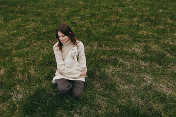 Naklejka premium A woman kneels on lush grass, cradling a small goat kid in her arms in a rural setting. Bright natural light highlights a serene moment of connection, care, and everyday animal companionship.