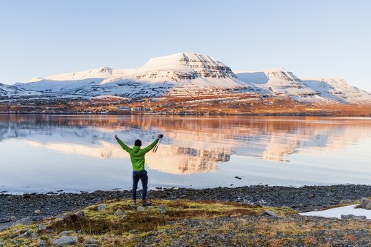 Man standing on the shore stretching his arms in the air, water reflection, snow-capped mountains in a fjord, Rey&eth;arfj&ouml;r&eth;ur, East Iceland, Iceland