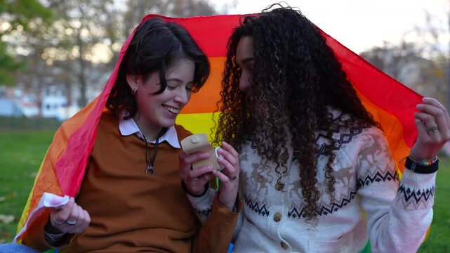 Two happy young women in love getting engaged under a rainbow flag in a park, showing their new ring