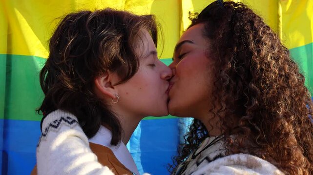 Young lesbian couple in love embracing and kissing against a rainbow flag background outdoors