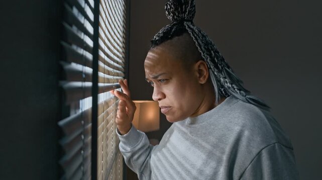 Side view chest up of Biracial woman with braided hair approaching apartment window and peeking through blinds while observing outside environment indoors
