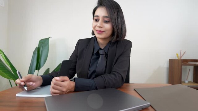 Closeup of thoughtful young Indian businesswoman working in office sits on chair at workplace desk holding pen and diary thinking creative ideas.