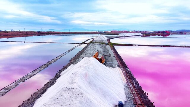Industrial view of a large white salt mound on a dirt road separating pink salt ponds with machinery in the distance. White salt heap on a pathway between pink lakes.
