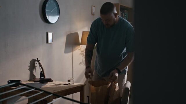 Tilt up of man wearing electronic ankle bracelet carrying paper bags and placing groceries on table in apartment during home confinement