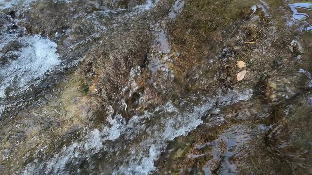 Slow motion of clear water rippling and flowing over natural brown rocks in a forest stream. Close up shot of water surface texture and small bubbles in 4K. Concept of purity, nature serenity, and hyd