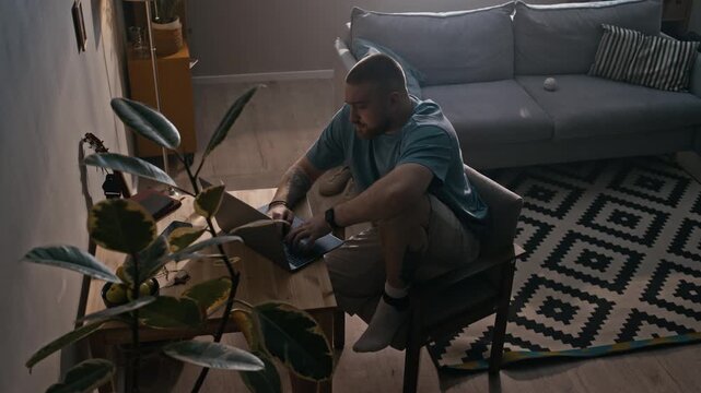 Eye level shot of Caucasian man under house arrest sitting at small desk and using laptop in apartment while wearing electronic ankle monitor