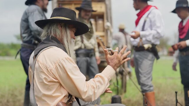 An old man sit showing off, brandishing a gun, in front of a cowboy caravan with day light.