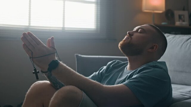 Tilt up of Caucasian man under house arrest sitting on apartment floor holding cross and praying while coping with isolation and emotional stress