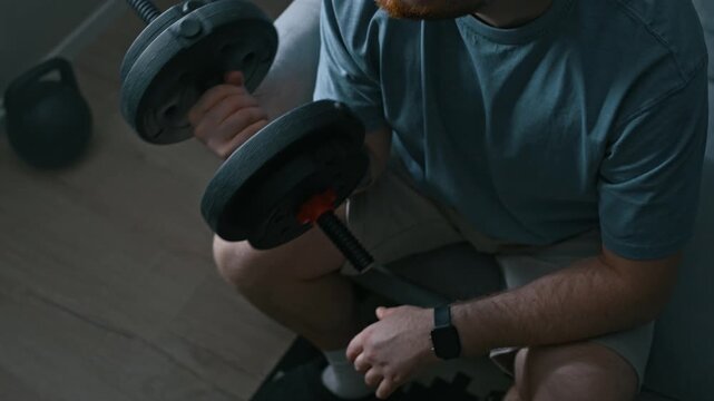 From above shot of tattooed man wearing electronic ankle bracelet lifting dumbbell while sitting on couch and exercising at home during confinement
