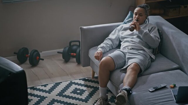 Eye level shot of Biracial woman with braided hair wearing ankle monitor lounging on couch drinking beer and watching television in apartment during house arrest