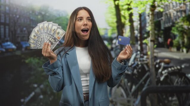 Woman holding a fan of dollars and a creditcard, hand raised in display on a street; financial independence excited.