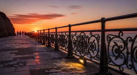 Obraz premium Wrought iron railing borders seaside promenade during vibrant sunset with figures walking in the distance