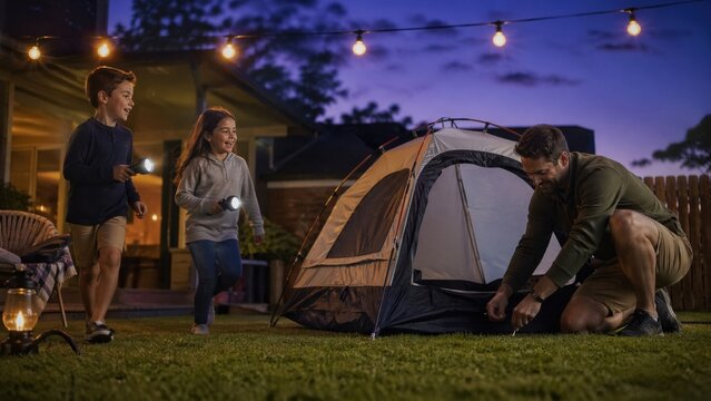 A smiling father pitches a small tent on a grassy lawn while his young son and daughter play with flashlights