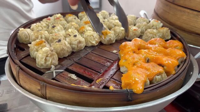 A bamboo steamer filled with two types of steamed dim sum, pale dumplings and bright orange ones topped with sauce. A hand uses metal tongs to pick one piece.