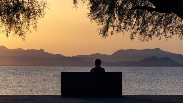 Mallorca seascape and landscape, with silhouetted and unrecognizable person sitting on a bench, looking at the mediterranean sea and Serra de Tramuntana in the distance.