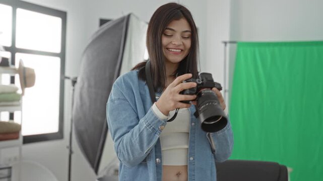 Young woman photographer adjusts a dslr camera and smiles while reviewing shots in a studio with green screen and softbox lighting; creative joy.