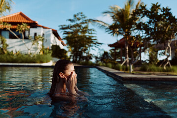 Fototapeta premium Young woman with wet hair covering her face while standing in an outdoor pool. Tropical trees and houses in the background. Concept of relaxation and lifestyle.
