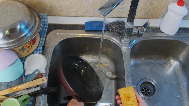 female hands washing pan in kitchen sink top view under running water, scrubbing cookware with sponge, removing grease and residue, household chore routine, daily cleaning activity