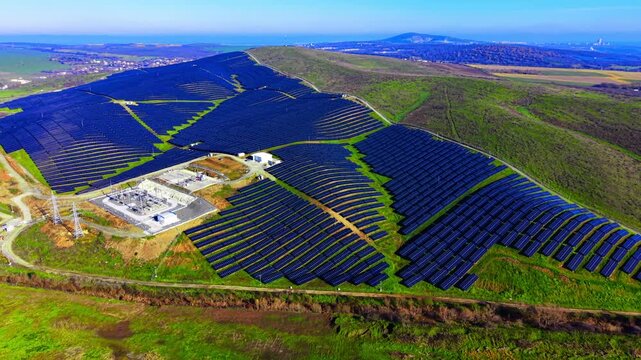Panoramic aerial view of a massive solar farm integrated into a hilly landscape with grid infrastructure. Industrial solar power plant with electrical substation on hills.