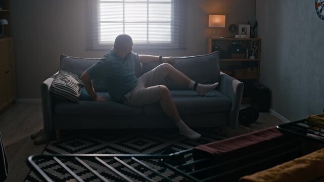 Wide shot of Caucasian man wearing electronic ankle bracelet getting up from couch in his apartment, looking frustrated, lonely, and emotionally exhausted during house arrest