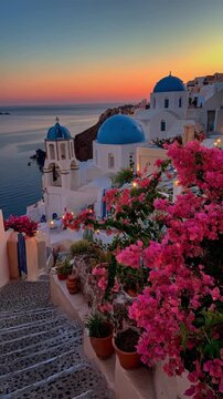Stunning sunset view of Oia village in Santorini with blue domed churches, white architecture, and pink bougainvillea flowers overlooking the Aegean Sea.