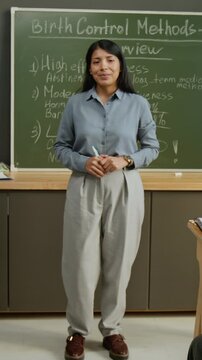 Full vertical dolly portrait of confident young Hispanic female teacher standing in front of blackboard with chalk notes on birth control methods during health education lesson, looking at camera