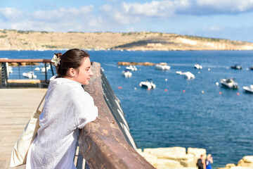 Fototapeta premium A young woman in a white shirt stands on a wooden pier looking at the blue sea with several small boats and a rocky coastline in the background.