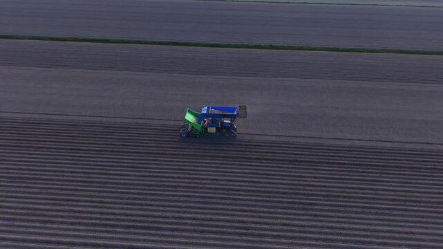 Aerial view of a blue agricultural machine planting seeds in a dark furrowed field under a dim sky in Middenbeemster, North Holland, Netherlands.