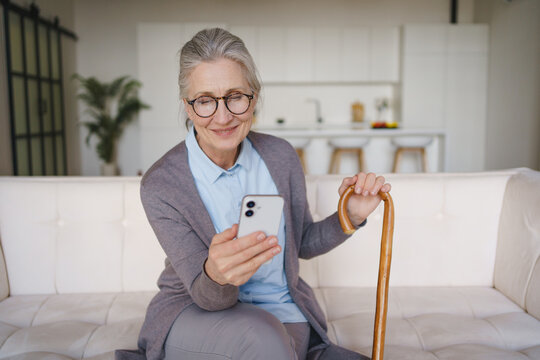 Senior woman with wooden cane using smartphone at home, elderly lifestyle with technology, online communication, independence, healthcare support, digital connection and modern aging concept