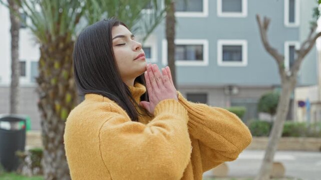 Young latina woman with hands pressed in prayer and eyes closed, wearing a yellow sweater on an urban street; serenity reflection prayer.
