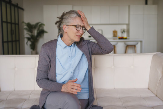 Senior woman with headache sitting at home, touching head, elderly lifestyle, pain, stress, tiredness, health problem, medical care and wellness concept