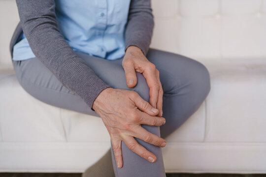 Close up of senior woman touching painful knee at home, elderly health problem, joint pain, arthritis, injury, healthcare, rehabilitation and pain relief concept