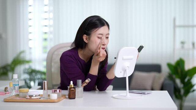 Asian women applying makeup using lighted mirror cosmetics beauty routine skincare eyebrow pencil grooming sitting home living room morning selfcare